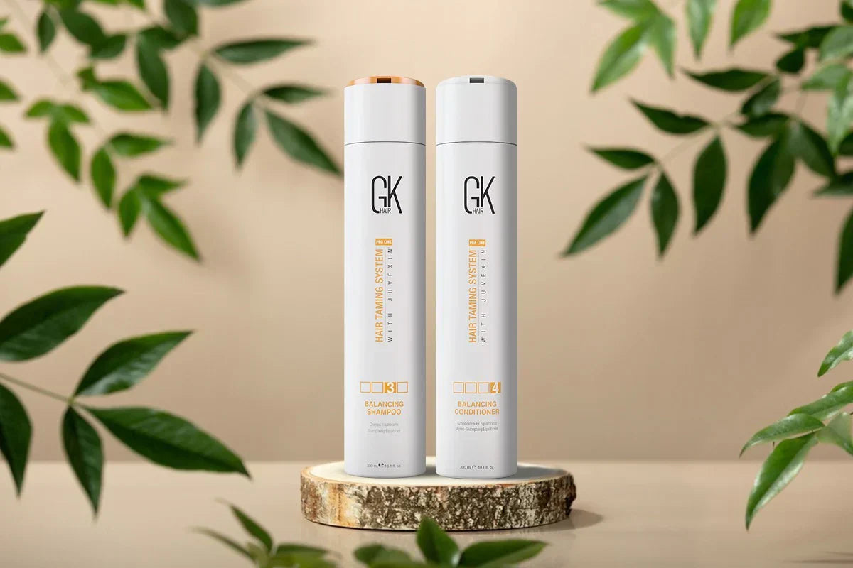 Two bottles of GK Hair Balancing Shampoo and Conditioner are displayed on a wooden slice, surrounded by green leaves on a soft background.